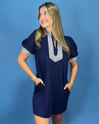 A woman modeling a navy blue short-sleeve mini dress with a striped V-neckline and striped puff sleeves, standing against a solid blue background.