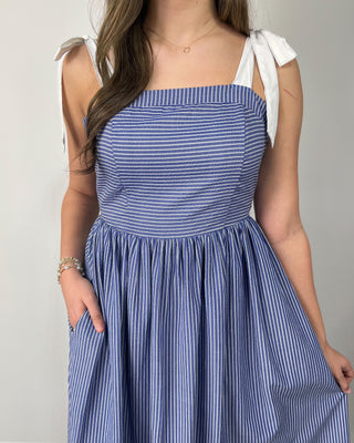 Model wearing a blue and white striped midi dress with bow tie shoulder straps, square neckline, gathered skirt, and side pockets, styled with nude heeled sandals.