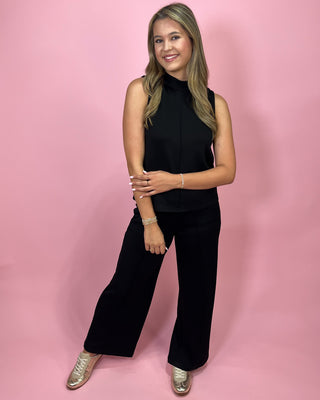 Woman wearing a black sleeveless top and black pants against a pink background