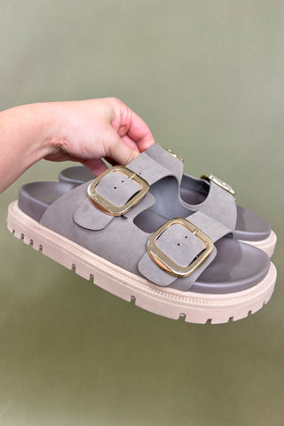 Gray sandals with gold buckles held by a hand against a green background