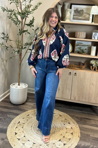 A smiling woman with long, wavy brown hair wears a navy blouse with bold red, white, and green floral embroidery, paired with high-waisted wide-leg jeans. She stands indoors on a round woven rug beside a potted plant and a wooden shelf decorated with artwork, candles, and small decor pieces.