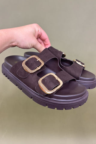Brown sandal with gold buckles held by a hand against a green background