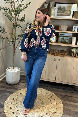A smiling woman with long, wavy brown hair wears a navy blouse with bold red, white, and green floral embroidery, paired with high-waisted wide-leg jeans. She stands indoors on a round woven rug beside a potted plant and a wooden shelf decorated with artwork, candles, and small decor pieces.