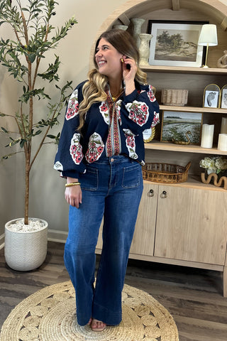 A smiling woman with long, wavy brown hair wears a navy blouse with bold red, white, and green floral embroidery, paired with high-waisted wide-leg jeans. She stands indoors on a round woven rug beside a potted plant and a wooden shelf decorated with artwork, candles, and small decor pieces.