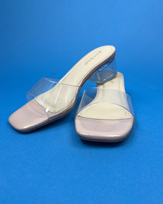 A neutral pair of slip-on heels with a clear heel and clear strap on a blue background, 