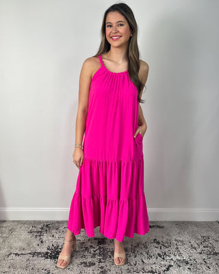 Model wearing a bright fuchsia tiered midi dress with braided halter neckline and flowy silhouette, styled with nude heeled sandals.