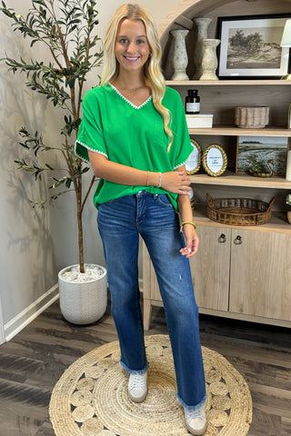 A smiling woman with long blonde hair wears a bright green V-neck top with white trim along the neckline and sleeves, paired with blue straight-leg jeans with frayed hems and white sneakers. She stands indoors on a woven circular rug beside a potted plant and a wooden shelf decorated with artwork, candles, and small decor pieces.