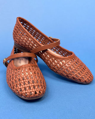 A pair of woven brown leather Mary Jane flats with openwork detailing and adjustable straps, displayed on a solid blue background.