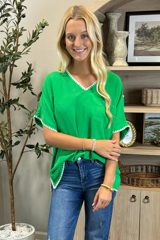 A smiling woman with long blonde hair wears a bright green V-neck top with white trim along the neckline and sleeves, paired with blue straight-leg jeans with frayed hems and white sneakers. She stands indoors on a woven circular rug beside a potted plant and a wooden shelf decorated with artwork, candles, and small decor pieces.