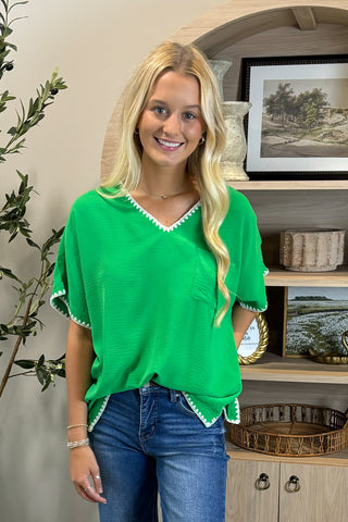 A smiling woman with long blonde hair wears a bright green V-neck top with white trim along the neckline and sleeves, paired with blue straight-leg jeans with frayed hems and white sneakers. She stands indoors on a woven circular rug beside a potted plant and a wooden shelf decorated with artwork, candles, and small decor pieces.