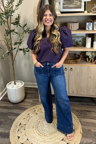 A smiling woman with long, wavy brown hair wears a short-sleeved deep purple puff-sleeve top paired with high-waisted wide-leg jeans with large front pockets. She stands indoors on a woven circular rug beside a potted plant and a wooden shelf decorated with candles, artwork, and small decor pieces.