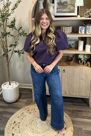 A smiling woman with long, wavy brown hair wears a short-sleeved deep purple puff-sleeve top paired with high-waisted wide-leg jeans with large front pockets. She stands indoors on a woven circular rug beside a potted plant and a wooden shelf decorated with candles, artwork, and small decor pieces.