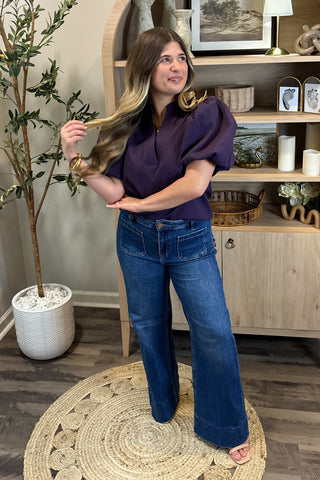 A smiling woman with long, wavy brown hair wears a short-sleeved deep purple puff-sleeve top paired with high-waisted wide-leg jeans with large front pockets. She stands indoors on a woven circular rug beside a potted plant and a wooden shelf decorated with candles, artwork, and small decor pieces.