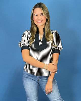 A woman modeling a black and white horizontally striped short-sleeve top with a solid black V-neckline detail, paired with light blue jeans, set against a solid blue studio background.