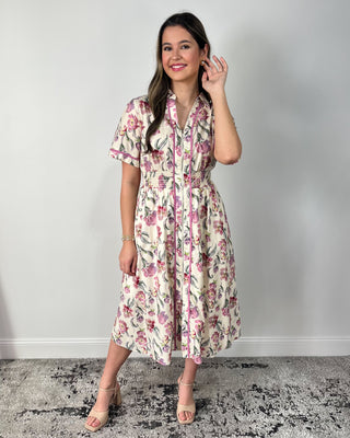 Model wearing a cream floral midi dress with short sleeves, collared neckline, zip-front detail, smocked waist, and side pockets, paired with nude heeled sandals.