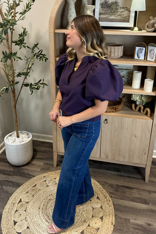 A smiling woman with long, wavy brown hair wears a short-sleeved deep purple puff-sleeve top paired with high-waisted wide-leg jeans with large front pockets. She stands indoors on a woven circular rug beside a potted plant and a wooden shelf decorated with candles, artwork, and small decor pieces.