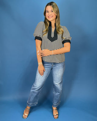 A woman modeling a black and white striped short-sleeve top with a black ruffled collar, paired with light-wash blue jeans and metallic sandals, standing against a solid blue studio backdrop.