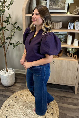 A smiling woman with long, wavy brown hair wears a short-sleeved deep purple puff-sleeve top paired with high-waisted wide-leg jeans with large front pockets. She stands indoors on a woven circular rug beside a potted plant and a wooden shelf decorated with candles, artwork, and small decor pieces.