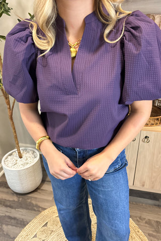 A smiling woman with long, wavy brown hair wears a short-sleeved deep purple puff-sleeve top paired with high-waisted wide-leg jeans with large front pockets. She stands indoors on a woven circular rug beside a potted plant and a wooden shelf decorated with candles, artwork, and small decor pieces.