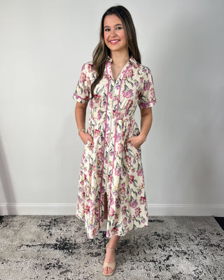 Model wearing a cream floral midi dress with short sleeves, collared neckline, zip-front detail, smocked waist, and side pockets, paired with nude heeled sandals.