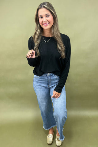 Woman wearing a black long-sleeve top and blue jeans against a green background