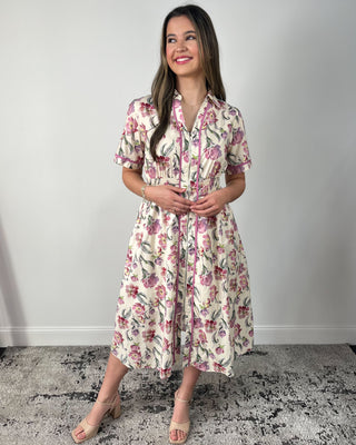 Model wearing a cream floral midi dress with short sleeves, collared neckline, zip-front detail, smocked waist, and side pockets, paired with nude heeled sandals.