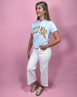 Woman wearing a light blue t-shirt with gold text and white pants on a pink background