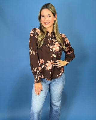 A woman modeling a brown long-sleeve blouse with a pink and beige floral pattern, paired with light blue jeans, standing against a solid blue studio background.