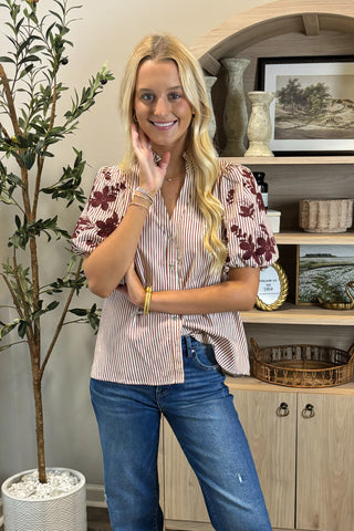 Woman in a striped shirt and jeans standing in a room with a plant and shelves.