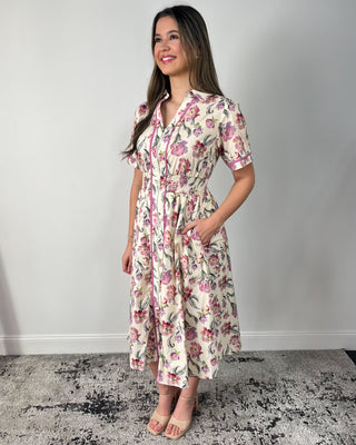 Model wearing a cream floral midi dress with short sleeves, collared neckline, zip-front detail, smocked waist, and side pockets, paired with nude heeled sandals.