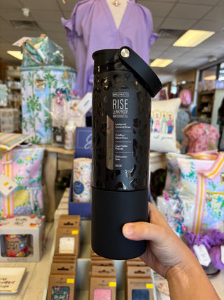 Hand holding a black mug with 'Rise' text in a store setting with various products and people in the background.