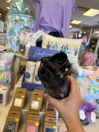 Hand holding a black mug with 'Rise' text in a store setting with various products and people in the background.