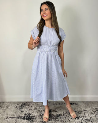 Smiling woman posing in front of a neutral backdrop wearing a breezy blue-and-white pinstripe midi dress featuring cap sleeves, a high round neckline, and a comfortable smocked waistband that creates a flattering fit-and-flare silhouette, styled with nude block-heel sandals and simple gold jewelry for an easy, polished look.