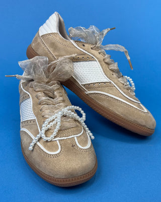 A pair of beige suede sneakers with white mesh panels, white laces decorated with pearl-like beads, and clear plastic wrapping around the laces, set against a solid blue background.