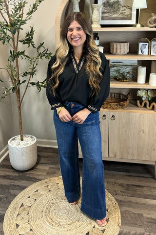 Woman in black top and blue jeans standing in a room with a plant and shelves.
