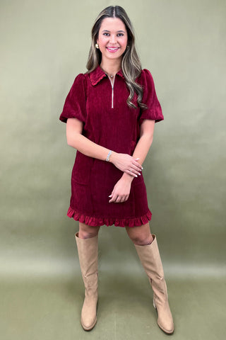 Woman wearing a burgundy dress with ruffled details and beige knee-high boots against a green background