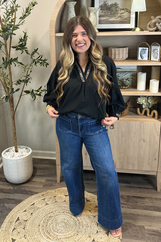 Woman wearing a black top and blue jeans standing in a room with a plant and shelves.