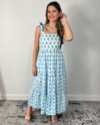 Smiling woman posing indoors wearing a breezy white maxi dress with dainty blue floral stripes, featuring a square neckline, bow-tie shoulder straps, and a tiered flowy skirt for the prettiest twirl-worthy movement, styled with nude block-heel sandals and simple gold jewelry for an effortlessly feminine spring-and-summer look.