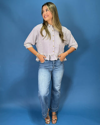 A woman modeling a white and blue vertically striped button-down blouse with a ruffled hem and gold buttons, paired with blue jeans and metallic sandals, standing against a solid blue studio backdrop.