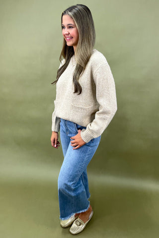 Woman wearing a beige sweater and blue jeans against a green background