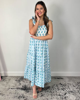 Smiling woman posing indoors wearing a breezy white maxi dress with dainty blue floral stripes, featuring a square neckline, bow-tie shoulder straps, and a tiered flowy skirt for the prettiest twirl-worthy movement, styled with nude block-heel sandals and simple gold jewelry for an effortlessly feminine spring-and-summer look.