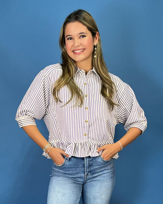A woman modeling a white button-down blouse with thin red vertical stripes, featuring puff sleeves, gold buttons, and a ruffled hem, paired with blue jeans against a solid blue background.
