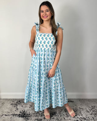 Smiling woman posing indoors wearing a breezy white maxi dress with dainty blue floral stripes, featuring a square neckline, bow-tie shoulder straps, and a tiered flowy skirt for the prettiest twirl-worthy movement, styled with nude block-heel sandals and simple gold jewelry for an effortlessly feminine spring-and-summer look.