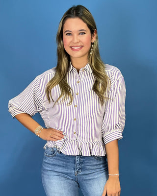 A woman modeling a white button-up blouse with thin purple vertical stripes, featuring puff sleeves and a ruffled hem, paired with blue jeans against a solid blue background.