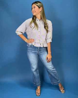 A woman modeling a white and red vertically striped button-down shirt with rolled sleeves and a ruffled hem, paired with blue jeans and metallic sandals, against a solid blue studio backdrop.