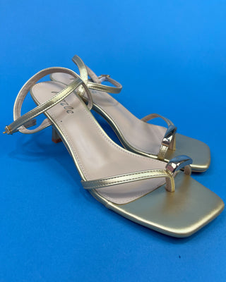 A pair of metallic gold strappy heeled sandals with square toes, shown on a solid blue background.