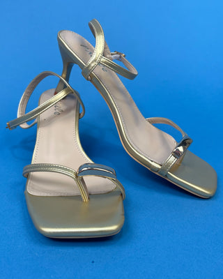 A pair of metallic gold strappy high-heeled sandals with square toes, shown against a solid blue background.
