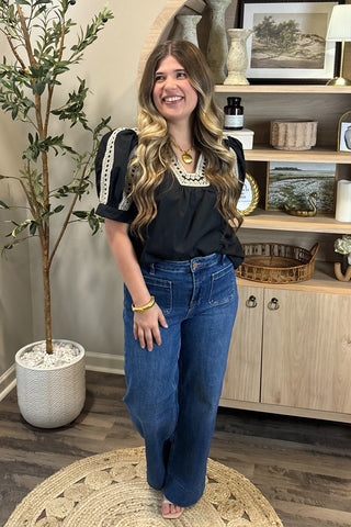 Woman in black top and blue jeans standing in a room with a plant and wooden cabinet.