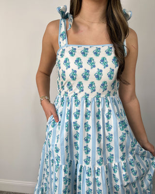 Smiling woman posing indoors wearing a breezy white maxi dress with dainty blue floral stripes, featuring a square neckline, bow-tie shoulder straps, and a tiered flowy skirt for the prettiest twirl-worthy movement, styled with nude block-heel sandals and simple gold jewelry for an effortlessly feminine spring-and-summer look.