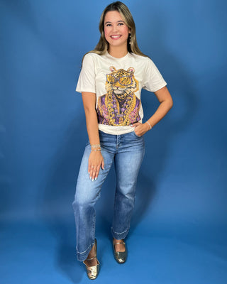 A woman modeling a cream-colored graphic t-shirt with a large gold and purple tiger print, paired with blue jeans and metallic gold flats, standing against a solid blue studio backdrop.